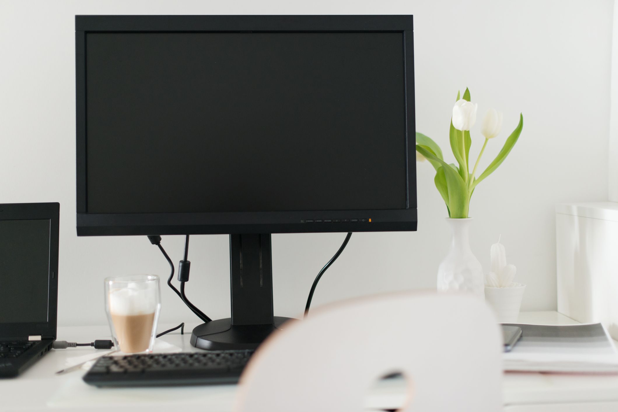 Монитор ПК на белом столе рядом с ноутбуком, белая ваза с цветами в нем и кофе
