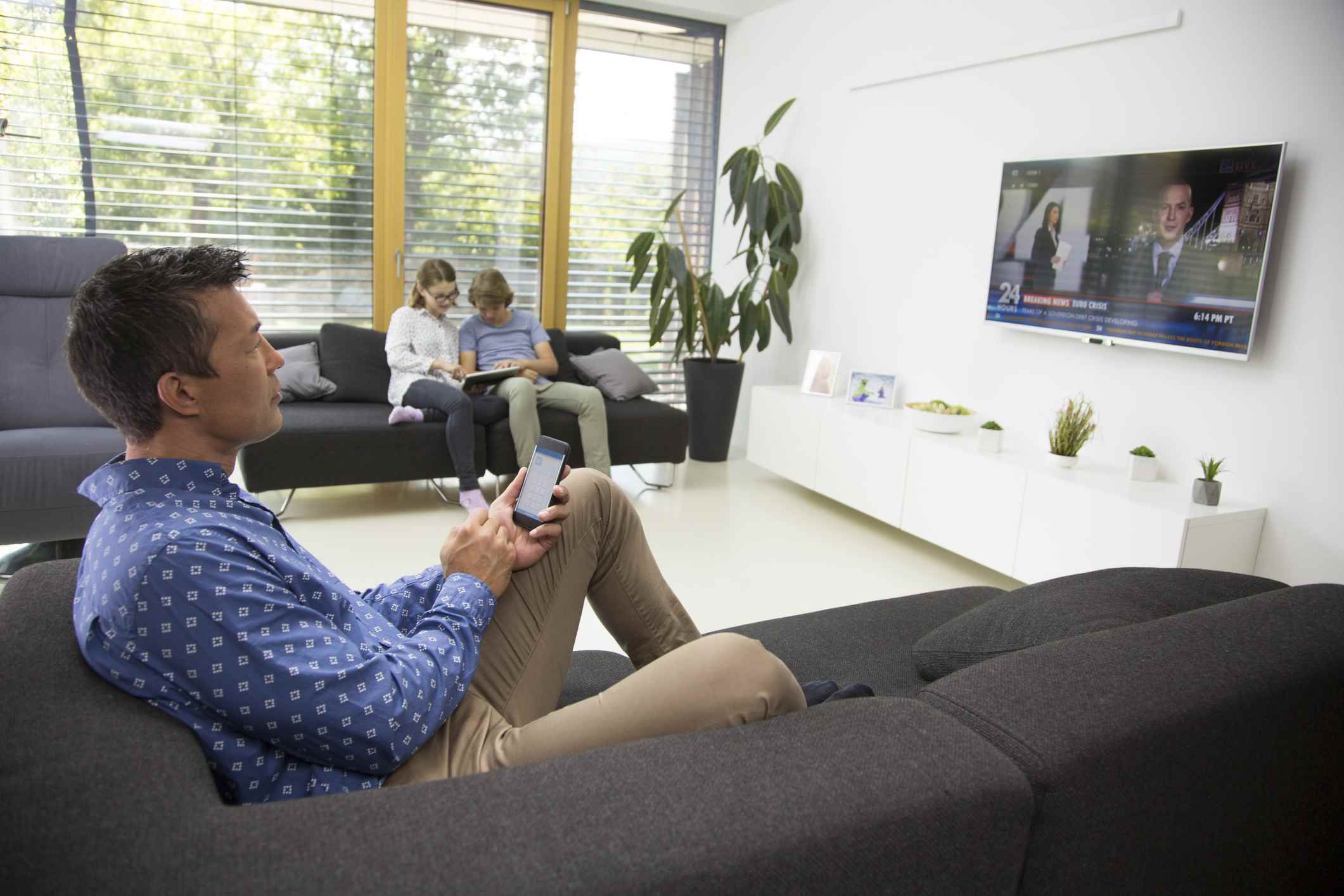Зрелый человек сидя на софе и контролируя smartphone пока смотрящ ТВ, детей используя цифровую таблетку в предпосылке.
